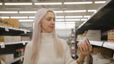 medium shot of house wife selecting canned food from supermarket shelf, tilts tin to examine expiry date printed on bottom under bright aisle lighting, weighing quality before placing into trolley