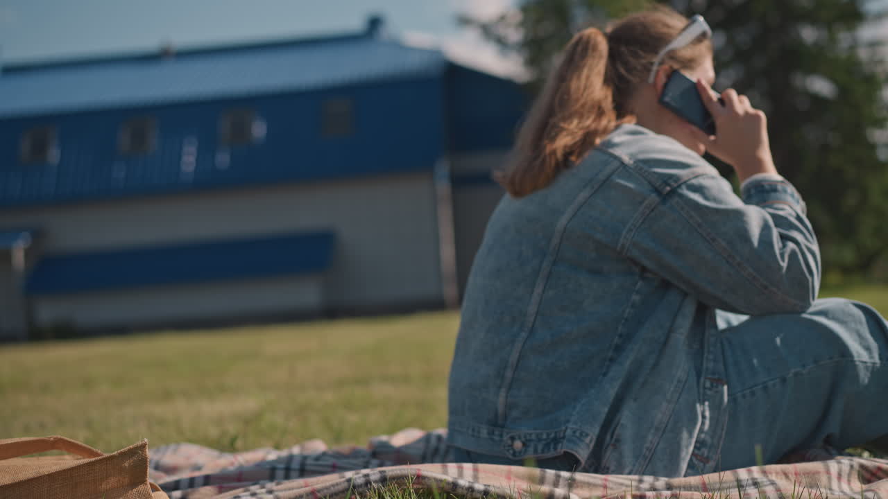 Donna caucasica seduta sull'erba che parla al telefono, che indossa una giacca di jeans con una borsa di paglia e una coperta nelle vicinanze, un edificio urbano sullo sfondo, luce solare del tardo pomeriggio, studentessa che pianifica scene, freelance.