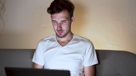 Ein-Junger-Mann-Mit-Stoppeln-Und-Moderner-Frisur,-Trägt-Ein-Weißes-T-Shirt,-Sitzt-Zu-Hause-Auf-Einem-Sofa,-Plaudert-Mit-Jemandem-Auf-Einem-Laptop,-Lächelt-Und-Lacht,-Amüsiert-über-Die-Antwort,-Statische-Nahaufnahme-4k