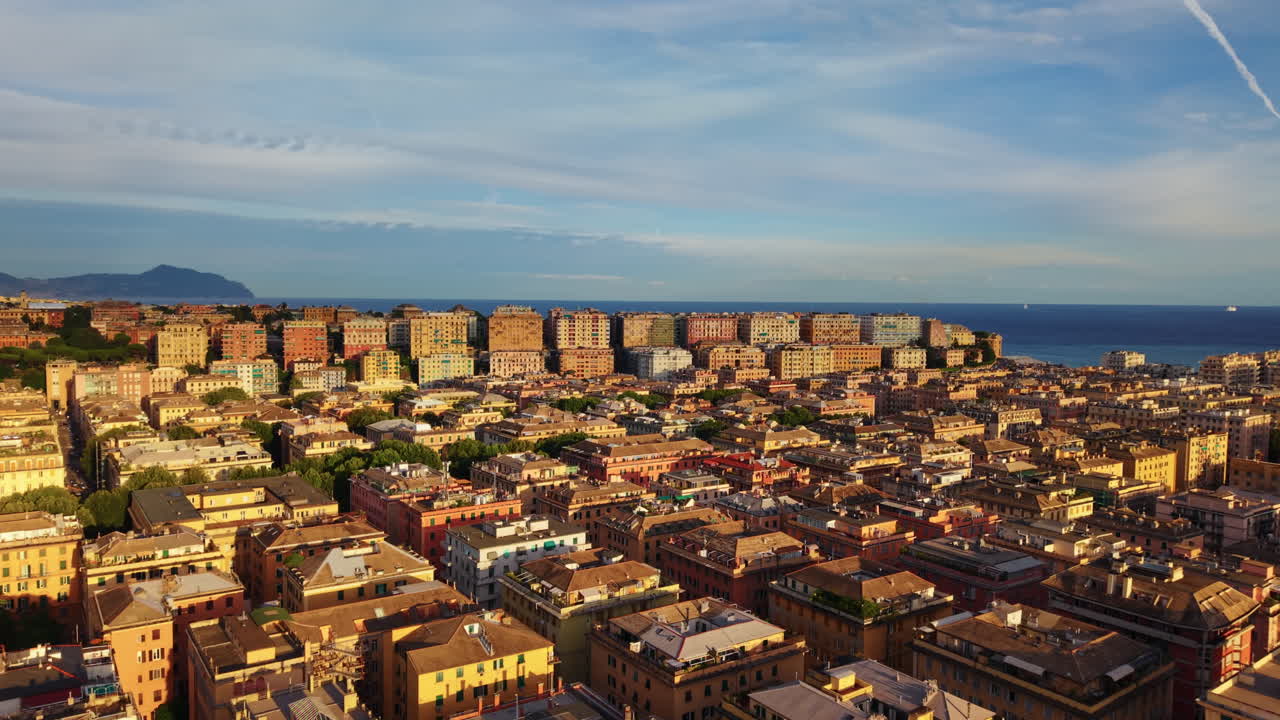 Drone advances above Genoa at sunset, showing uniform rooftops, higher buildings on a hill, the sea and mountains in the background