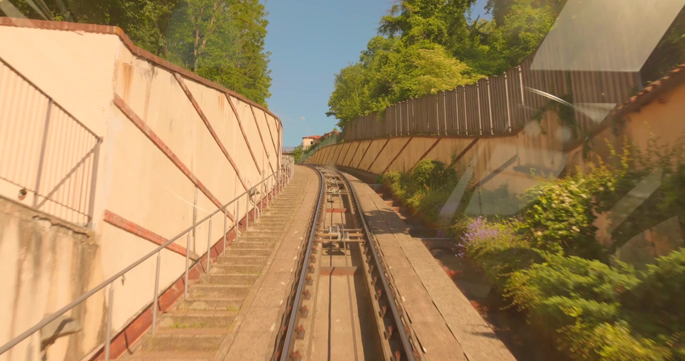 Pemandangan dari dalam kereta api Funicular Mondovi yang curam mendaki bukit di antara dinding penahan dan vegetasi hijau pada hari yang cerah, Mondovi, Piedmont, Italia