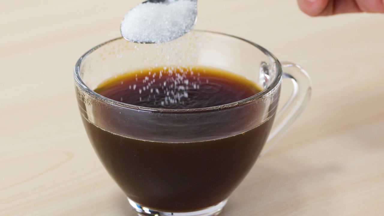 Primer plano de azúcar blanca cayendo de una cuchara en una taza de cristal con café negro