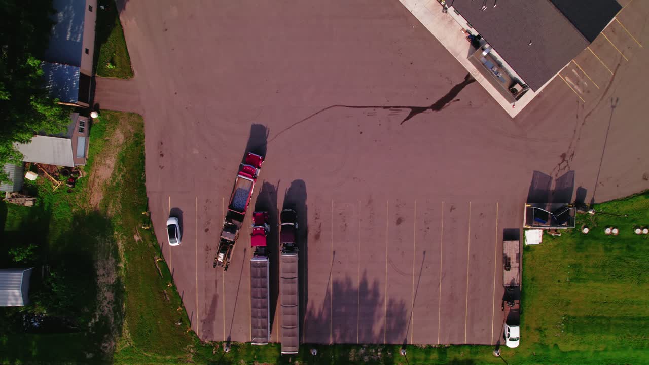 Top-down aerial view of a red semi-truck backing up in a rural parking lot beside other parked hopper grain trucks. Minnesota, USA