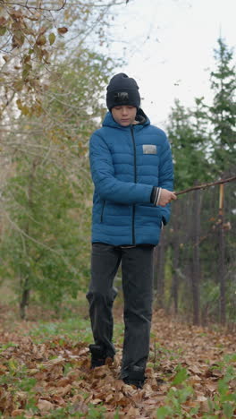 joven con chaqueta azul caminando por el sendero de otoño con un palo en la mano esparciendo las hojas secas en el suelo, rodeado de hojas secas, interactuando lúdicamente con la naturaleza en el bosque