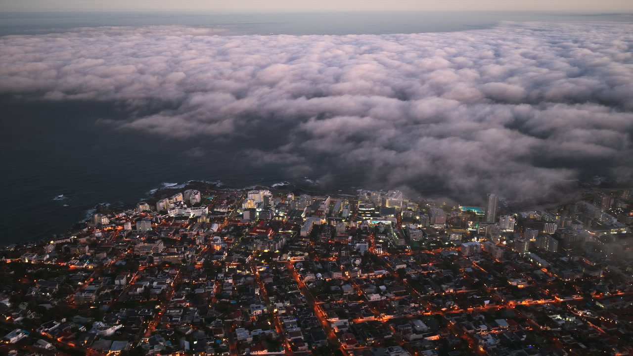 A stunning aerial view of Cape Town at dusk, where glowing city lights meet the vast ocean, as a dramatic blanket of clouds rolls in from the horizon.