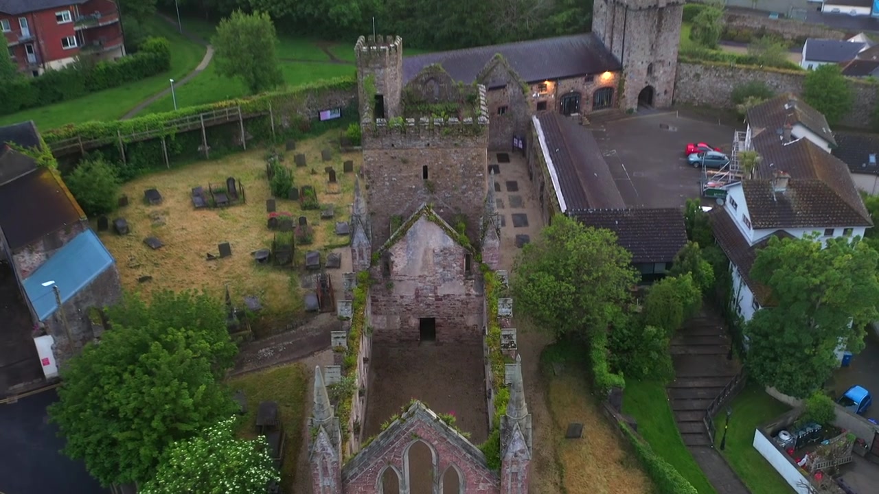Descending aerial reveals Selskar Abbey from wide establishing perspective through dynamic rotation around 12th-century tower base showcasing graveyard headstones and gothic architectural detail
