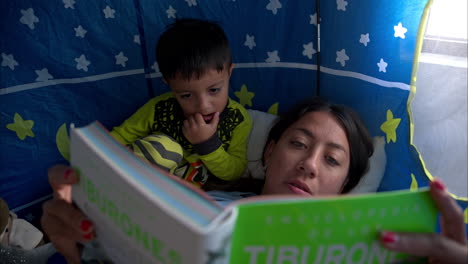 primer plano de una madre mexicana latina morena leyendo un libro a su hijo acostado dentro de su tienda de castillo teniendo un momento juntos