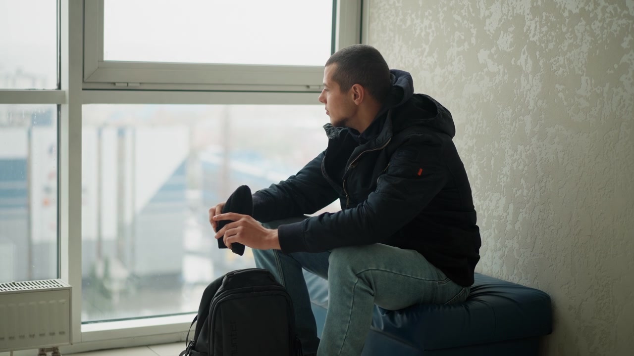 Close up of young man sitting by window holding and adjusting his cap while looking outside. Man wears dark jacket with hood and light blue jeans black backpack rests on floor beside him