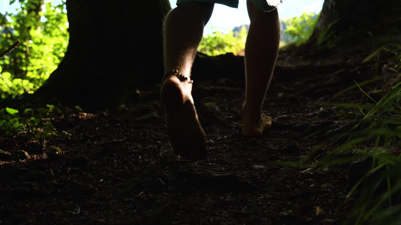 Closeup of mans feet walking barefoot in slow motion on a path out of a dark forrest towards a bright green meadow in with sunshine