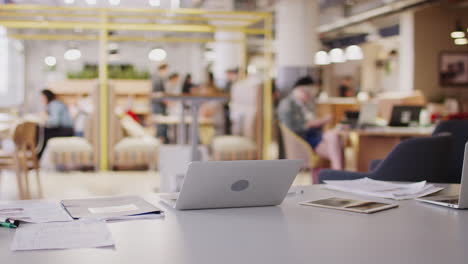 laptop computer and paperwork on a desk in a busy open plan creative office space, selective focus