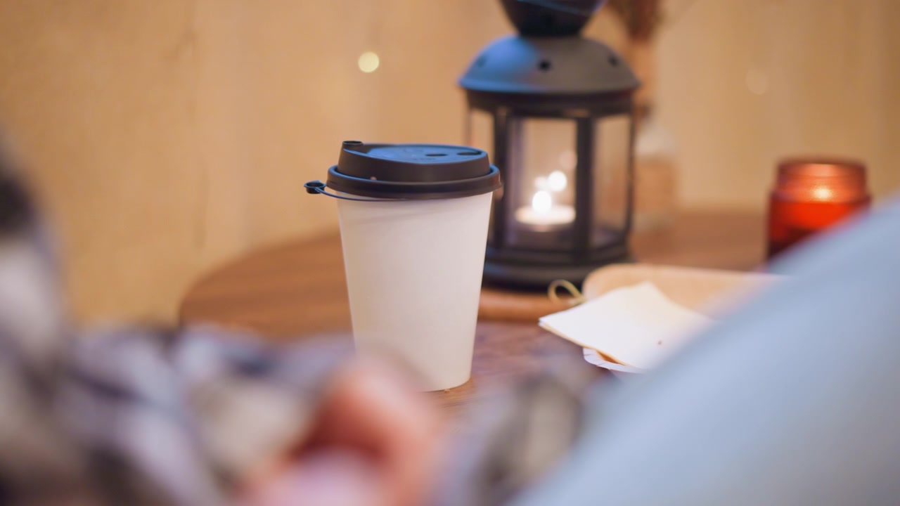 Primo piano di una tazza di caffè usa e getta bianca con coperchio nero su un tavolo di legno. Lo sfondo presenta una lanterna accesa e una candela; l'illuminazione calda e soffusa crea un'atmosfera accogliente. Persona sfocata con camicia a quadri.