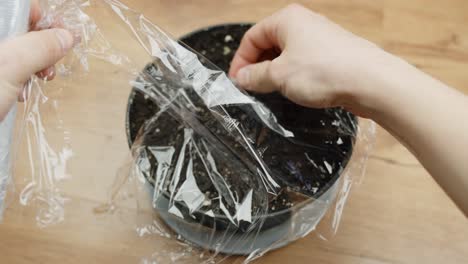 A top-down view of hands gently covering a pot of soil and seeds with cling film, creating a greenhouse effect for growth.