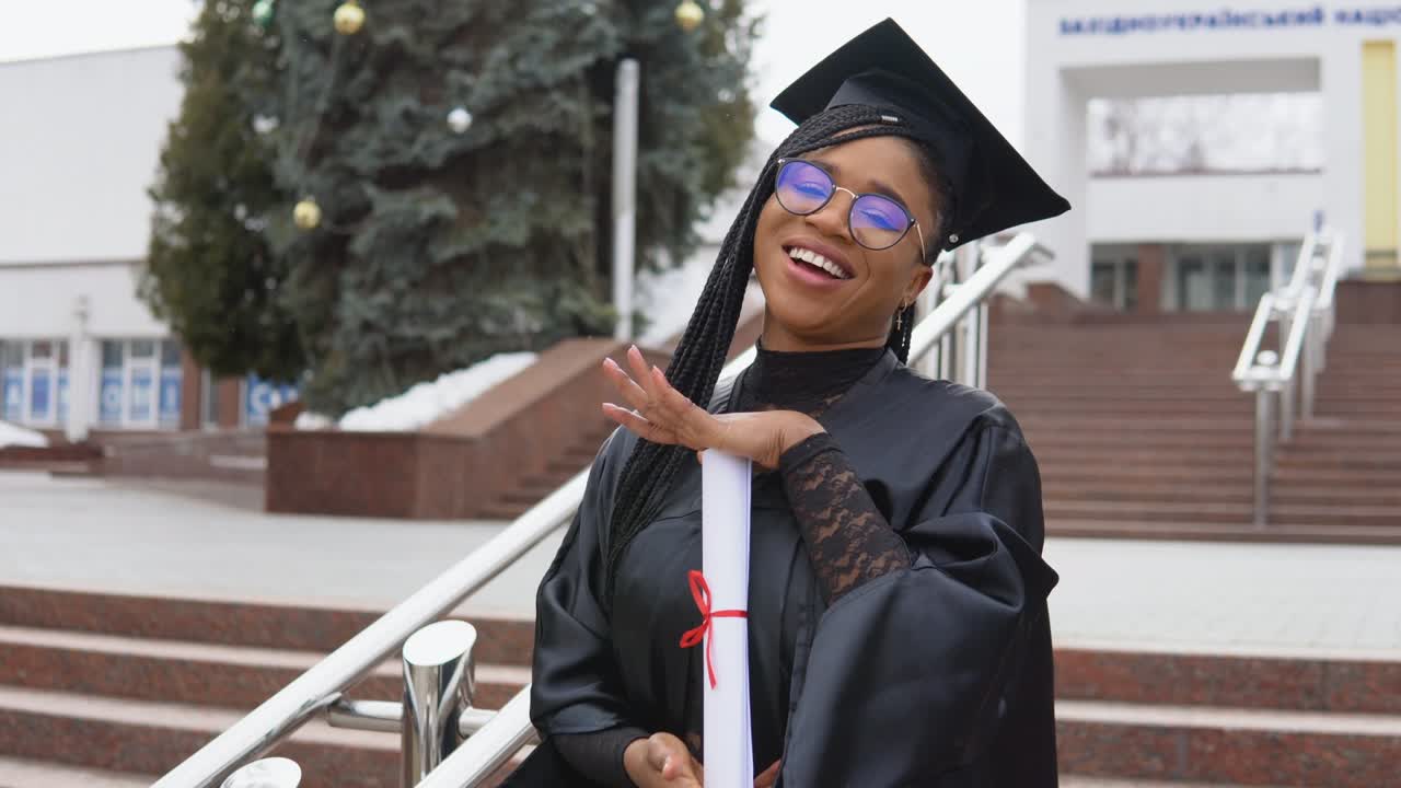 una mujer joven en la universidad en el manto del maestro sostiene un diploma y mira a la cámara. retrato con las escaleras frente a la entrada central de la universidad en fondo
