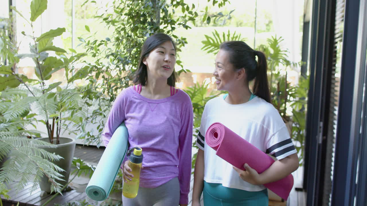 Happy asian female friends with exercise mats walking and smiling on sunny terrace, slow motion