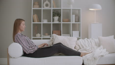 boring-young-woman-is-chatting-in-internet-by-laptop-resting-at-home-at-weekend-lying-on-couch-in-living-room