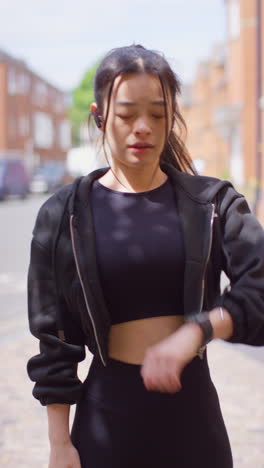 video vertical de una mujer joven haciendo ejercicio corriendo en una calle urbana usando auriculares inalámbricos comprobando un reloj inteligente 1