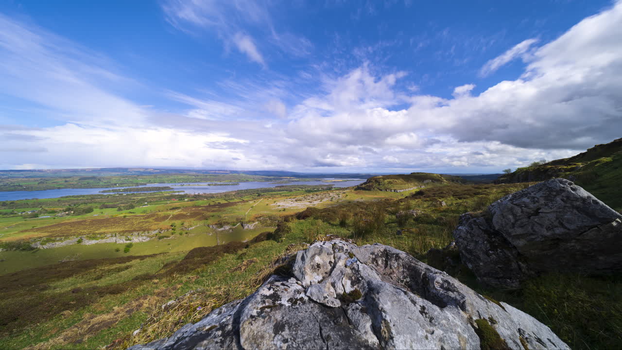 Timelapse av et landlig landskap med fjellformasjon i forgrunnen og åser og innsjø i det fjerne på en solrik og skyet dag, sett fra Carrowkeel i grevskapet Sligo i Irland