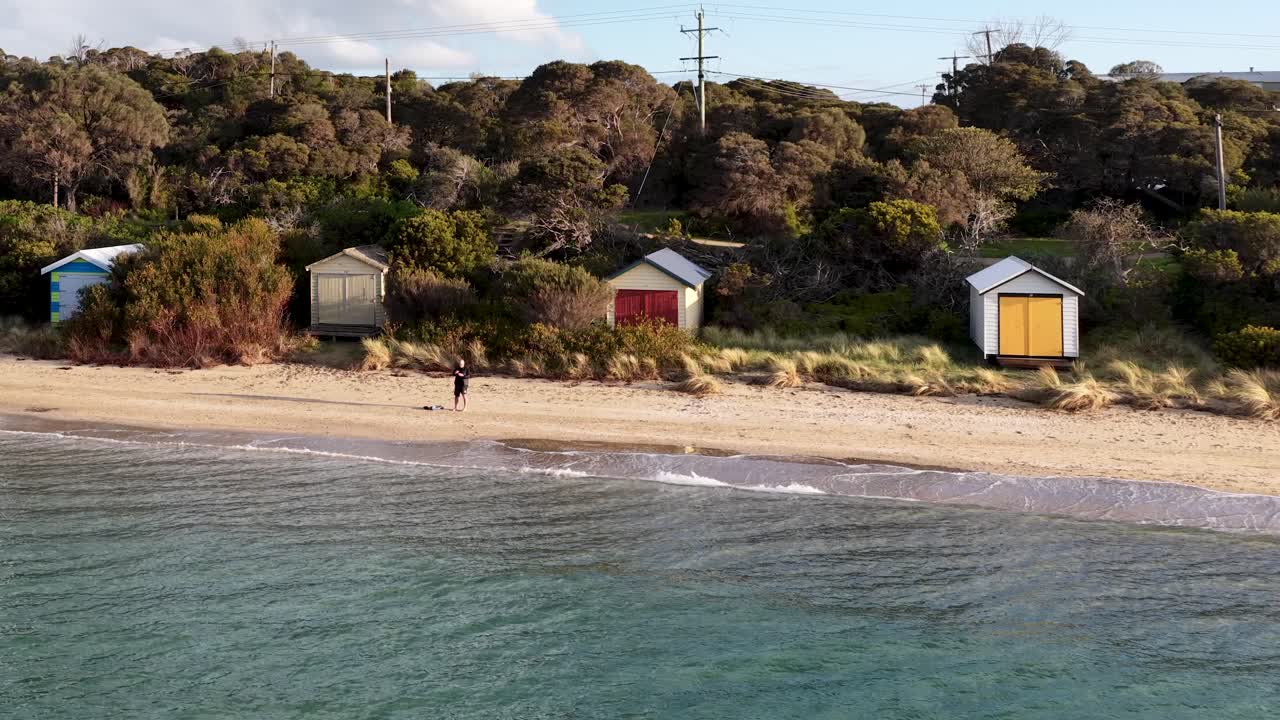 Drone glides over colorful beach huts, tranquil shoreline, and coastal vegetation in soft evening light