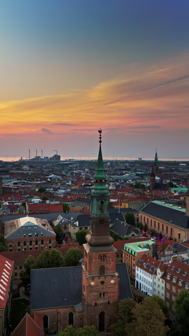 Aerial drone view of St. Nicholas Church, with its green copper spire rising above the city's red rooftops at sunset in Copenhagen, Denmark. Vertical