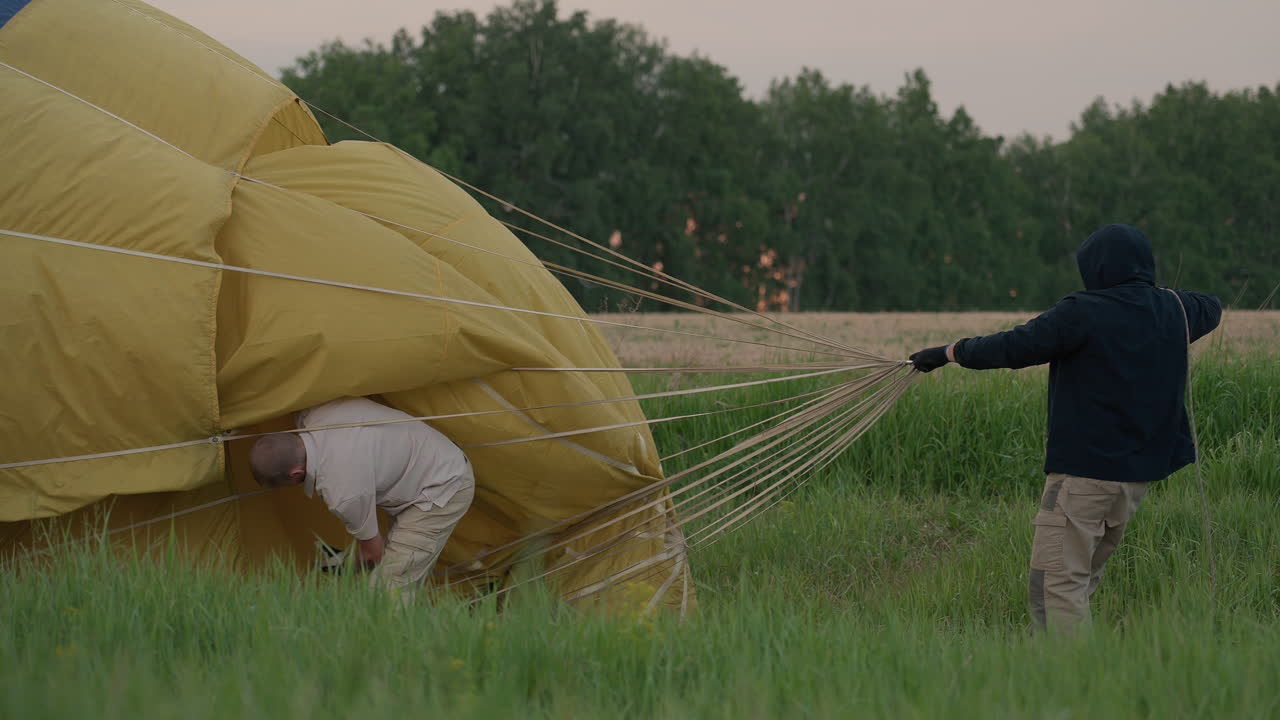 person in dark jacket pulling collapsed hot air balloon envelope across green grass field under moody sunset sky preparing balloon packing operation in rural evening landscape setting