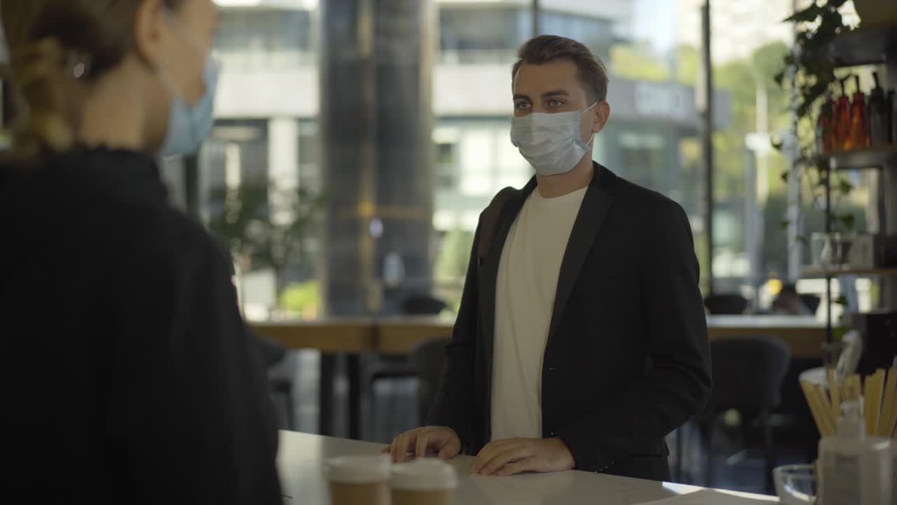 joven empresario positivo con máscara facial de covid-19 recibiendo café para llevar en el mostrador de la cafetería y saliendo. retrato de un cliente satisfecho comprando una bebida refrescante por la mañana en la pandemia de coronavirus.