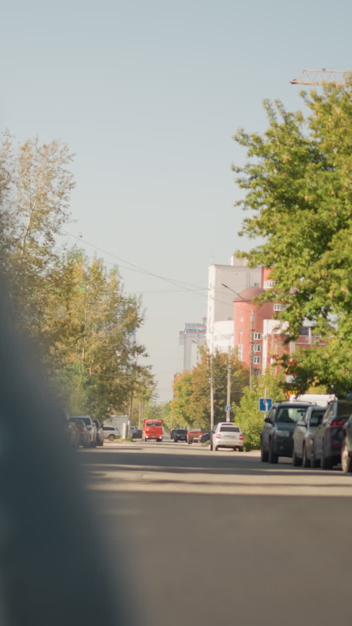 햇빛이 비치는 교외 거리, 주차된 자동차, 낮은 앵글의 도로에서 보이는 세단, 잎이 무성한 나무, 파스텔 색조의 상점 전면, 정오의 햇빛이 드리우는 긴 그림자, 꾸준한 교통 흐름 등이 포함되어 있어요.