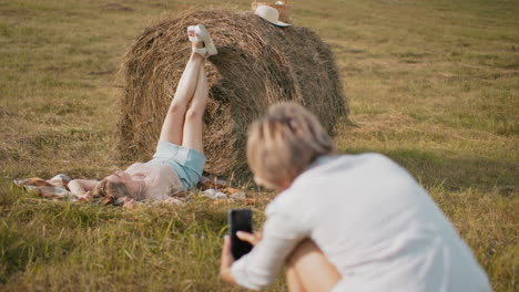 photographer capturing artistic shot of woman lying on hay bale in rural setting, model poses with legs up while photographer focuses in foreground, natural outdoor photography in warm countryside