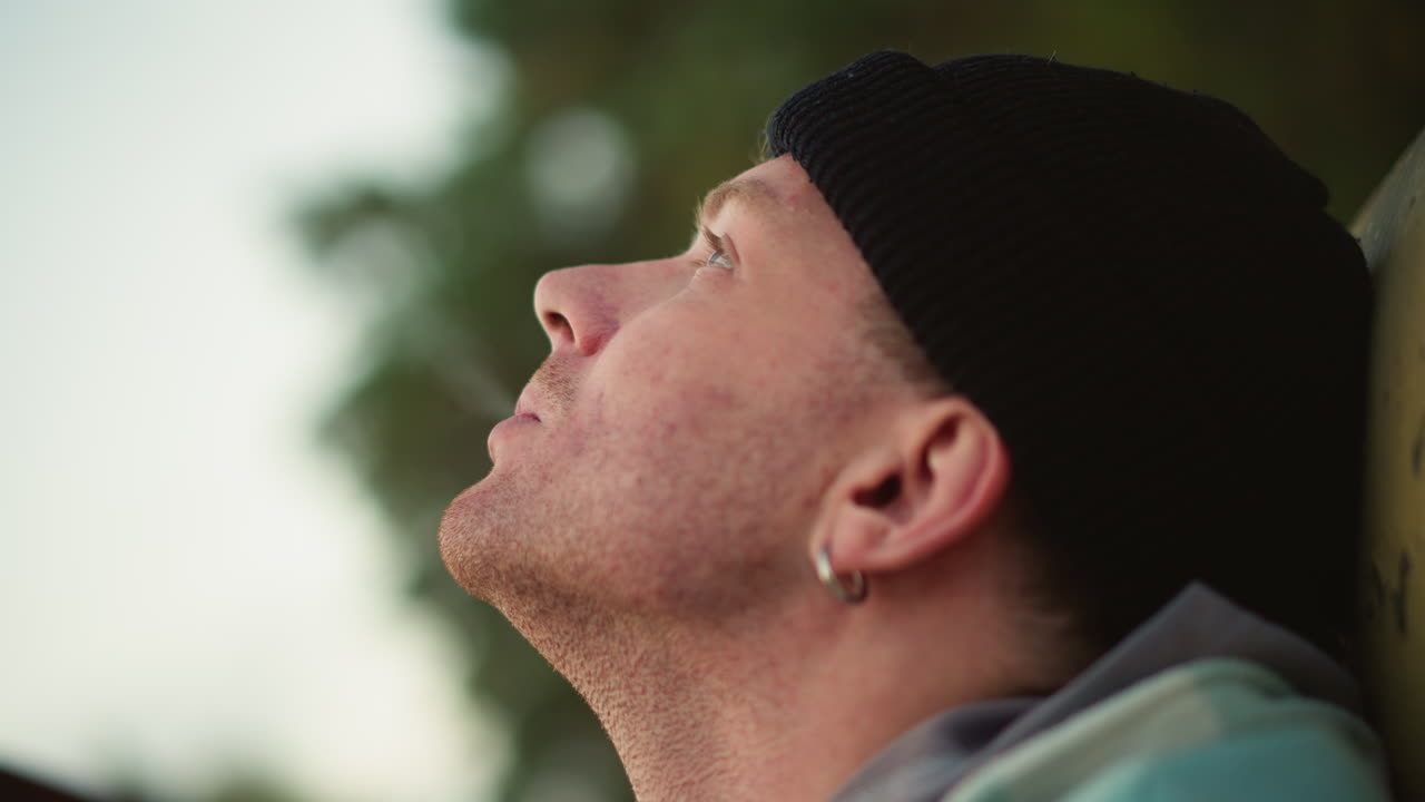 white man smoking cigarette outdoors leaning against railing, beanie and earring visible, rings on fingers, closeup profile shot, soft bokeh foliage background, dusk light, pensive expression