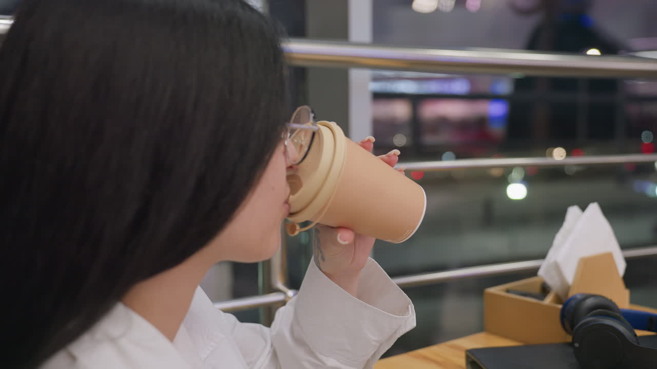 close up of woman in glasses enjoying coffee while holding phone, seated beside wooden table with night city traffic seen through glass panel, soft bokeh lights in background