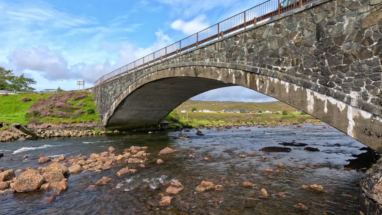 A steady camera captures a stone arch bridge spanning a shallow stream with rocks, lush green hills, and bright daylight on the Isle of Skye