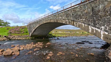 a steady camera captures a stone arch bridge spanning a shallow stream with rocks, lush green hills, and bright daylight on the isle of skye