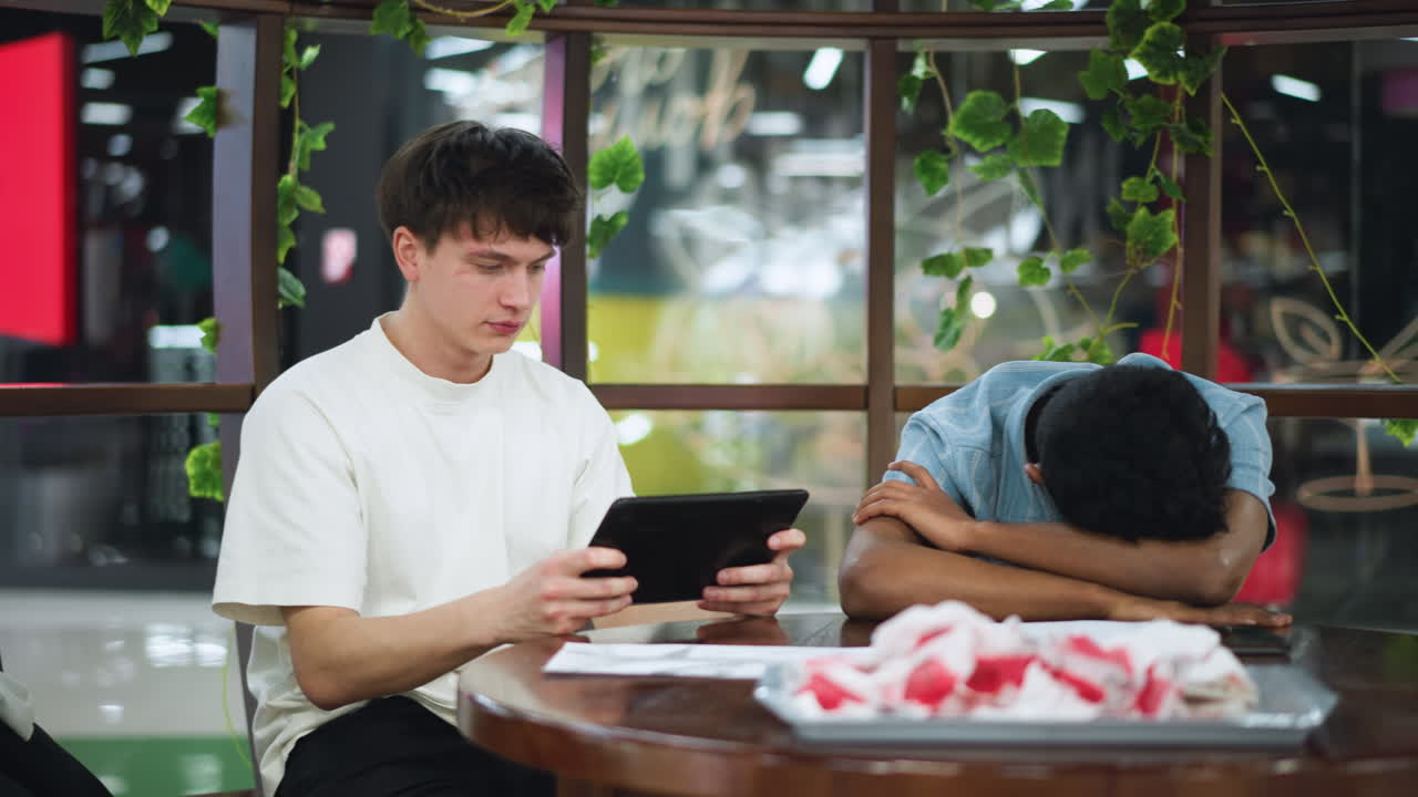 Very fair skinned lad wakes up at desk holding tablet and smartphone while friend rests head on table in relaxed mood inside modern classroom under soft morning light with subtle shadows around
