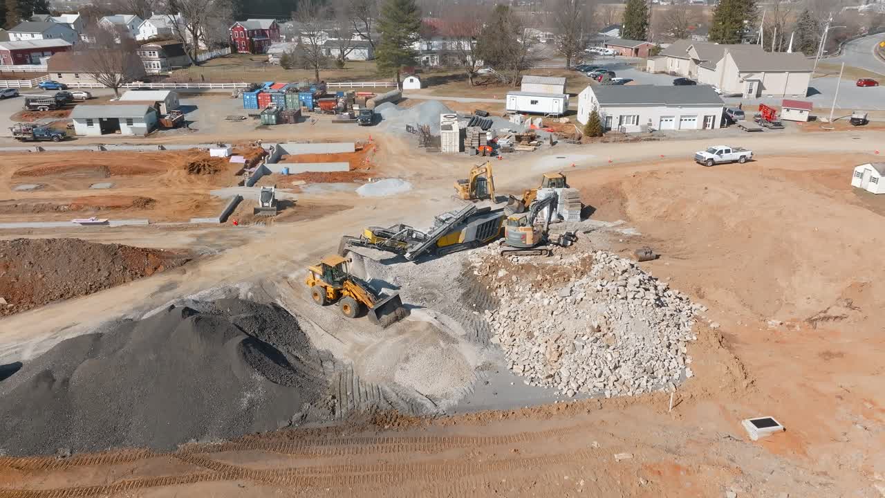 Heavy machinery operates at a construction site, moving rocks and gravel. Workers are busy sorting materials as the site is prepared for future development. Dust and equipment fill the area.