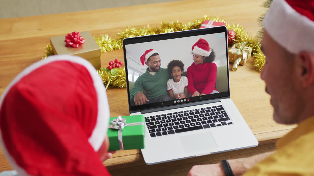 padre y hijo caucásicos con sombreros de santa usando una computadora portátil para una videollamada de navidad con la familia en la pantalla
