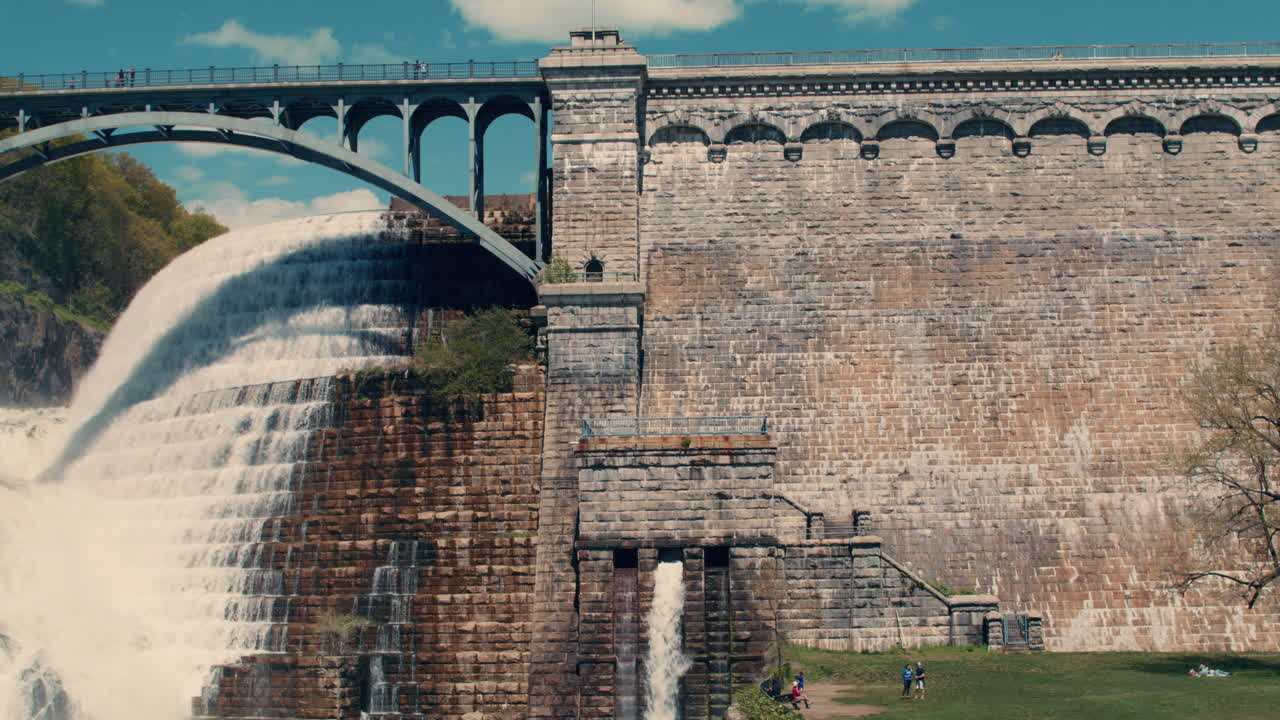 medium shot of New Croton Dam bridge and wall with stepped spillway, waterfall, and lawn visible with blue sky. slow motion 40 fps