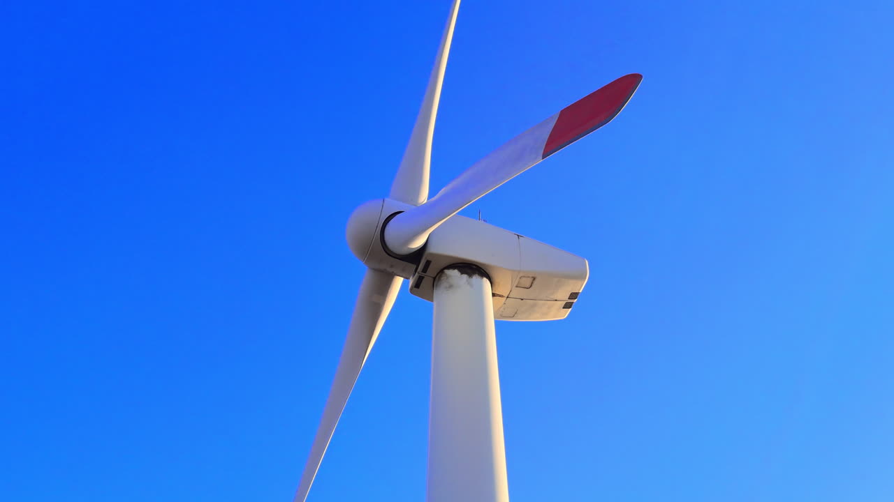 Góndola blanca de un aerogenerador y tres palas giratorias contra un fondo de cielo azul sólido a la luz del día. Primer plano de un aerogenerador. Vista aérea.