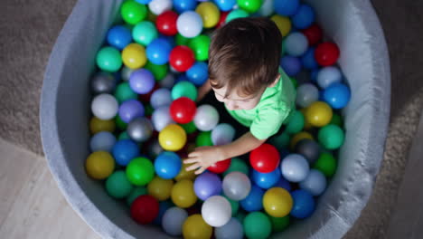 little toddler lies on the balls in the dry pool. baby boy raises head looking into camera and smiling. top view.