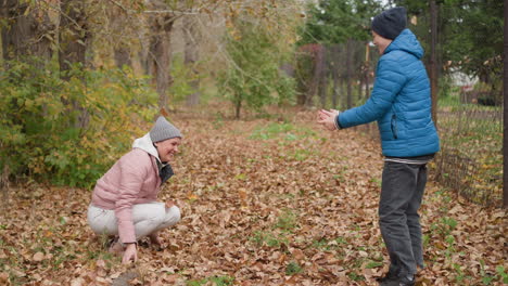 mother and son playing outdoors, surrounded by autumn leaves, as she sprays dry foliage in the air, both smiling and enjoying the moment, with green and autumn scenery in the background