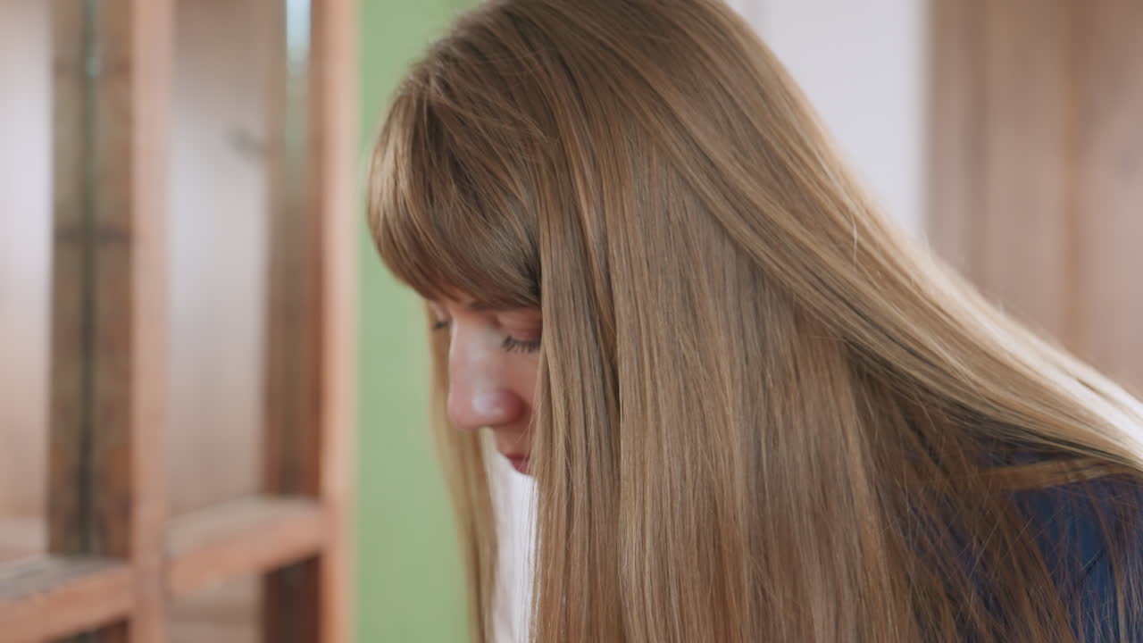 Specialist with long straight hair stands near mirror, reaching into bag with focused expression, soft indoor light casting gentle shadows across face, wood frame reflections enhancing quiet moment