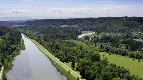 Aerial-view-at-a-little-boat-coming-down-the-isar-river-framed-by-green-colored-summer-trees---natural-scene-of-the-beautiful-bavarian-landscape-next-to-the-alps-mountains-in-the-background