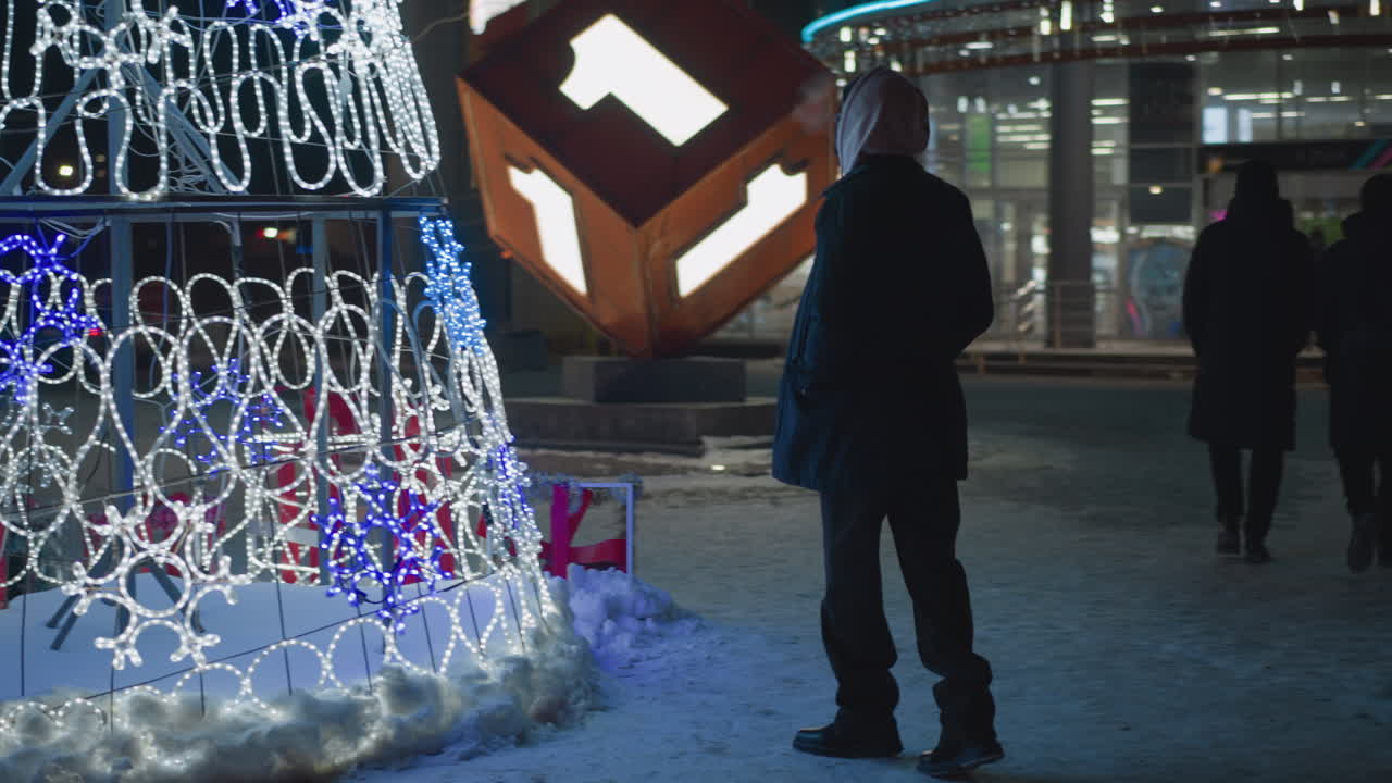 A person in dark winter coat stands facing brightly lit festive Christmas decorations in snow-covered urban area at night, surrounded by people in winter clothes walking in cold weather