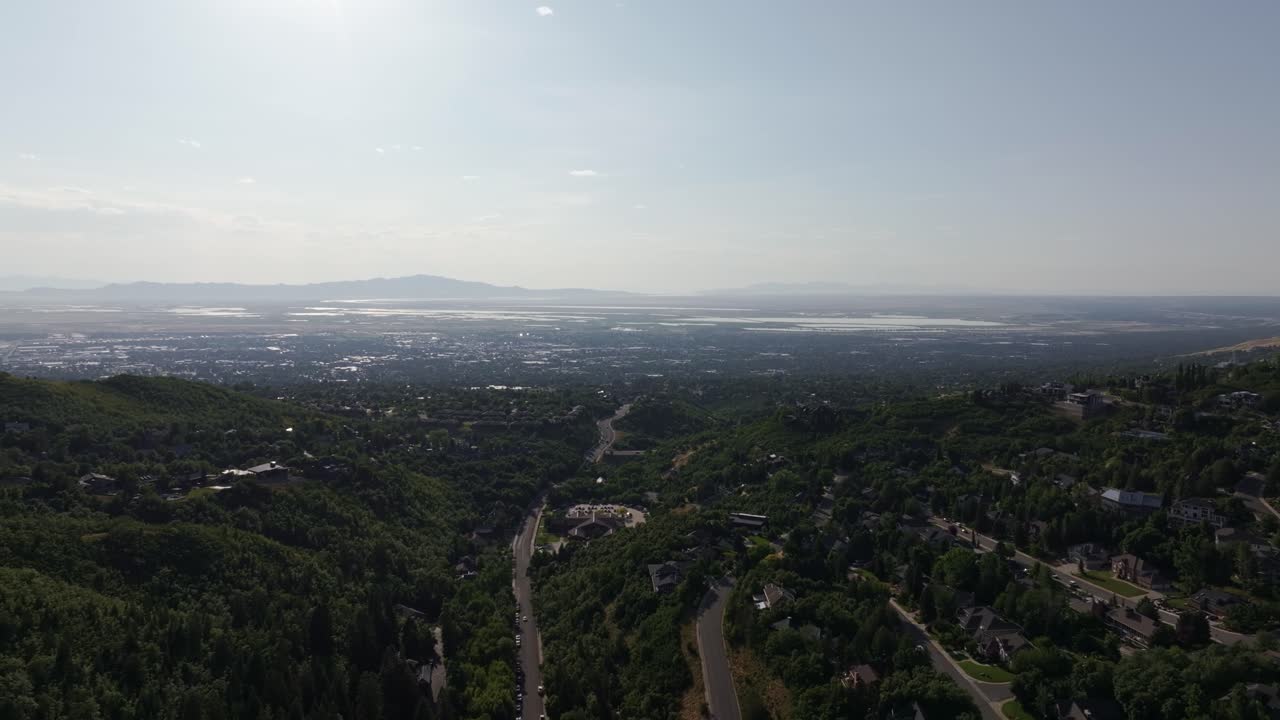A wide aerial drone dolly out trucking left shot from Bountiful Canyon showing a road, forest, valley homes, cityscape, mountains, and the Great Salt Lake on a clear summer day
