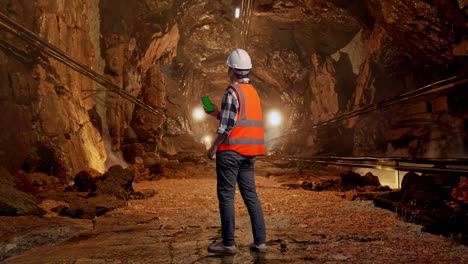 full body back view of asian male engineer with safety helmet working on a green screen smartphone and looking around while standing in underground mine tunnel
