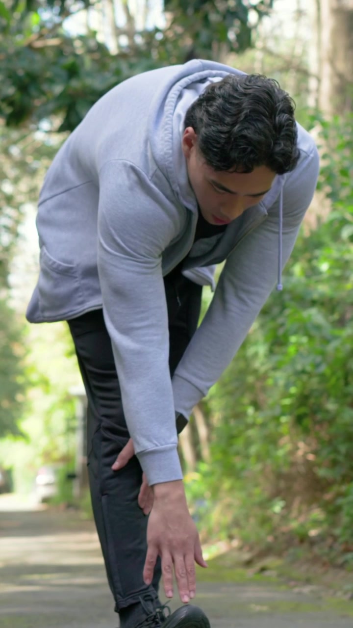 Vertical video: Feeling pain, bending adult man checking right ankle on park path, adjusting shoe