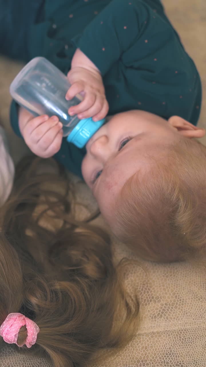 adorable little baby drinks water and elder sister in white t-shirt lies nearby on large bed in room view from above