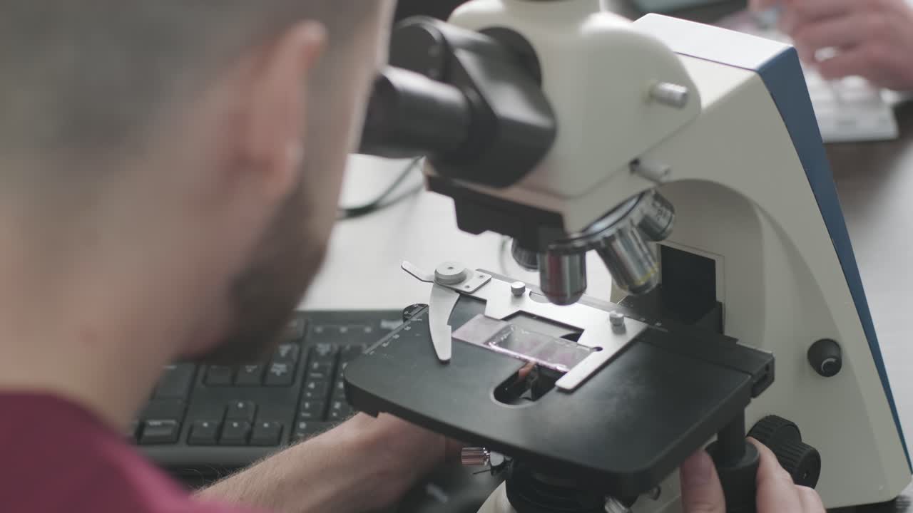 Male microbiologist, medical worker looks at blood sample through a microscope, makes analysis in laboratory. Laboratory assistant conducts histological research of biomaterial using modern microscope.