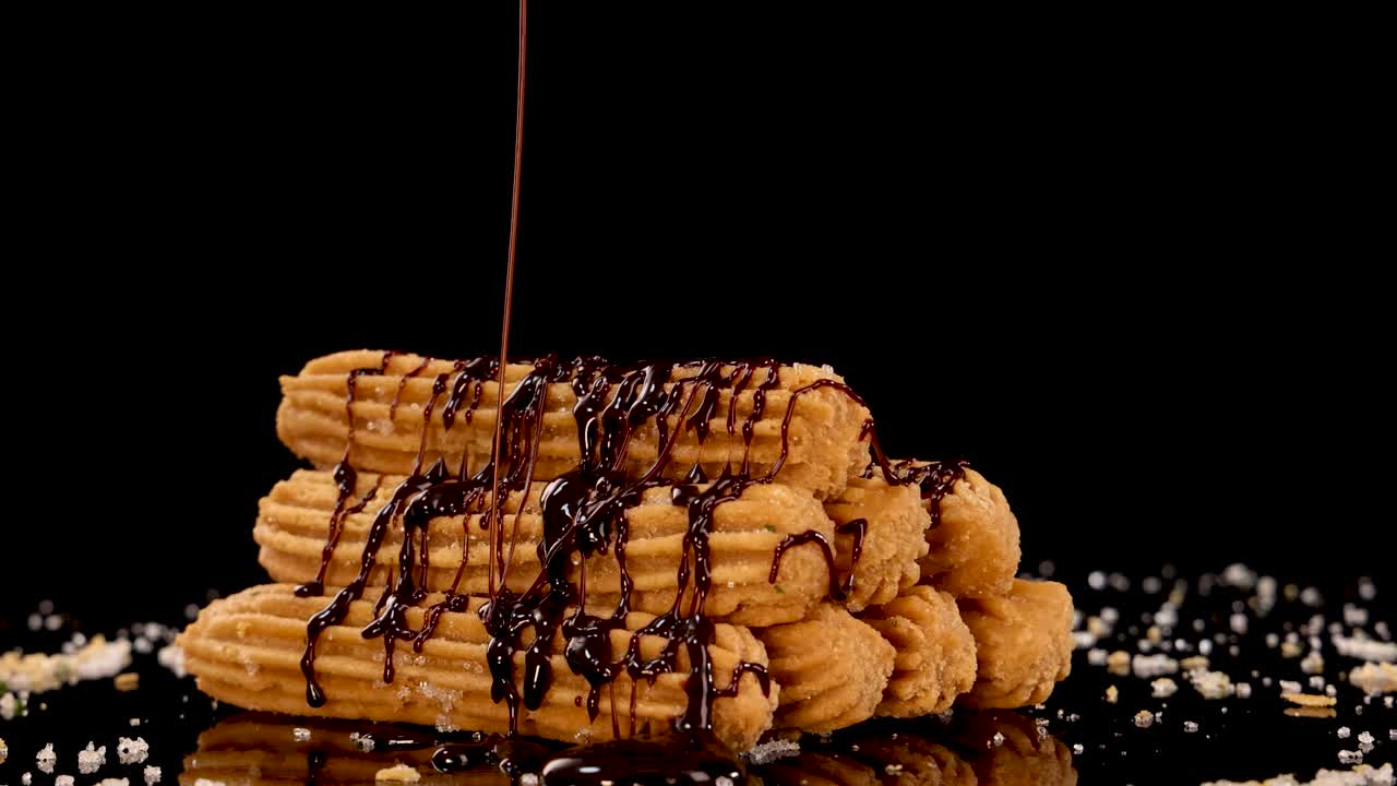 A stack of golden churros is slowly drizzled with rich chocolate sauce against a black background, with dramatic lighting and close-up composition
