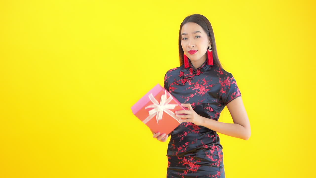 Asian model holding pink gift box with white lace ribbon and yellow background. Young woman in black and red dress smiles and admires present
