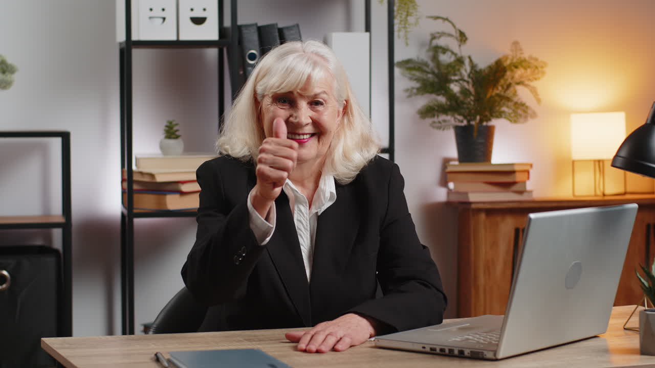 Happy caucasian elderly businesswoman with netbook showing thumbs up towards camera at home office