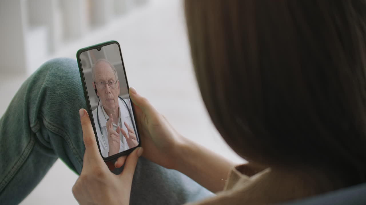 Female medical assistant wears white coat headset video calling distant patient on smartphone. Doctor talking to client using virtual chat telephone app. Telemedicine remote healthcare services